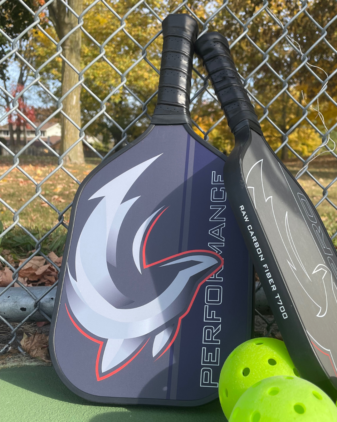 Slash Performance paddle leaning on a chain-link fence beside a second paddle and two yellow-green pickleballs