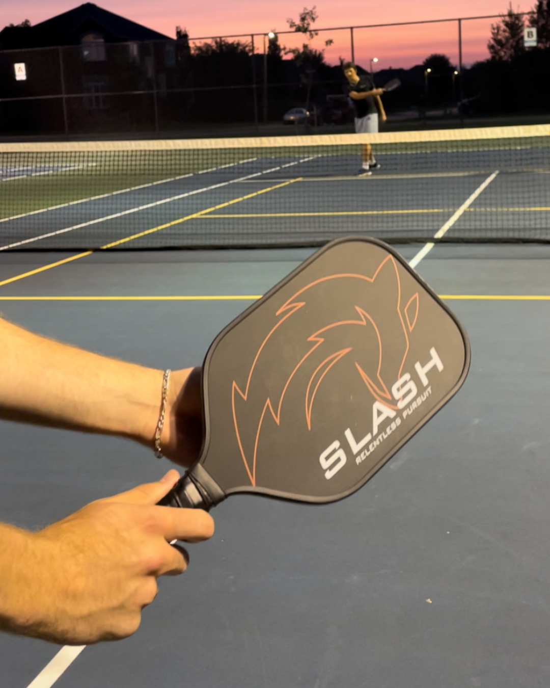 Hand gripping a Slash paddle in a serving position on an outdoor pickleball court at dusk